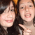 Two people with dark hair pose closely together, one wearing a black top and the other in light-colored clothing, both smiling against a blurred background.