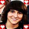 A person with shoulder-length brown hair and bangs smiles at the camera, framed by four red heart icons with white pixelated hearts in each corner.