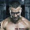 A shirtless man with short dark hair and visible ear piercings stands against a dark, blurred background with text reading "NO MERCY" at the bottom.