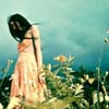 A woman in a flowing peach dress stands in a field of tall grass and flowers, her head bowed and dark hair obscuring her face against a blurred blue sky background.