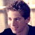 Pixelated static image of a person's head and shoulders with dark hair, light skin tone, and visible facial features including eyes, nose, mouth, and ear against a blurred background.