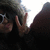 A person with light brown hair and white sunglasses makes a peace sign with their right hand against a bright background.