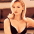 Static pixelated image of a woman with blonde hair wearing a black spaghetti-strap top, seated in an indoor setting with brown furniture and light walls.