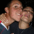 Two people in a close embrace, one with light brown hair and a dark shirt, the other with short light hair and a dark top, against a black background.