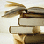Stack of three books with brown and beige spines, one book tilted upward showing a white page edge against light background.