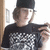 Pixelated image of a person wearing a black cap and dark shirt with white pattern, holding a phone near their face against a light background.