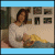 A woman in a white long-sleeve shirt sits on a bed with blue and yellow bedding, holding a yellow object while looking at the camera, then turning her head right and extending her arm forward.