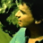 Pixelated static image shows a person with light skin and green hair wearing a light blue top against a dark background with green foliage.