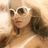 Pixelated image of a person with light skin tone wearing white sunglasses and dark hair, holding an object near the face against a blurred background.