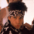 A person with dark curly hair wears a black and white patterned headband and dark clothing with white patterns, positioned against a light background with another figure partially visible behind.