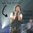 Pixelated image of a person holding a microphone with one hand near their mouth, standing against a dark background with light effects and another figure partially visible.