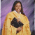 A pixelated figure in a yellow garment with dark hair against a purple background, holding an indistinct black object.