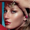 A close-up of a person with light skin, blue eyes, and red lips wearing a gold headpiece and pink nail polish, with a hand touching their forehead.