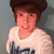 Pixelated image of a person with brown hair wearing a white shirt with dark blue text, set against a gray background.