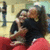 Two people in red and black clothing embrace in a gym setting with blurred background figures.