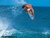 Pixelated surfer in mid-air above blue ocean waves, wearing dark shorts and light-colored top.