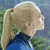 Pixelated profile view of a person with light brown hair in a high ponytail wearing a blue top against a blurred green and blue background.