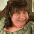 Pixelated image of a person with dark brown hair wearing a green patterned shirt, smiling with eyes closed against a blurred background.