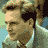 Pixelated static image of a man's face in profile with light brown hair, wearing a white shirt and green jacket against a blurred background.