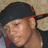 Pixelated close-up of a person wearing a dark cap with red trim, light skin tone, and visible facial features including eyes, nose, mouth, and ear.