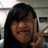 A pixelated close-up of a person with long dark hair, smiling broadly while holding an object near their face against a blurred background.
