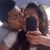 Two people pose closely together, one holding a dark rectangular object while the other leans in to kiss their cheek; background is light with blurred shapes.