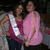 Two women in pink tops and silver sashes pose together, one with hand on hip, the other with arms crossed, in a dimly lit setting.