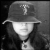 A person wearing a dark bucket hat with a white 'T' logo, long hair, and a circular pendant necklace appears in four grayscale showing text changes on the right