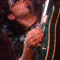 A person wearing dark sunglasses and a dark patterned shirt holds a green electric guitar with a white pickguard, performing under stage lighting.