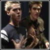 Two young men in dark blue shirts with white stripes perform synchronized hand gestures against a black background.