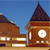 Pixelated image of a brown clock tower with a white clock face and dark roof against a blue sky, adjacent to a brown building with white windows.