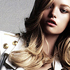 A close-up of a person with long wavy blonde hair wearing a white jacket with gold buttons against a light gray background.