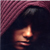 Pixelated profile view of a figure with dark hair under a maroon checkered hood, face illuminated by warm light against dark background.