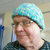 Pixelated portrait of a person wearing a light blue cap and dark clothing, with a blurred background showing indistinct shapes and colors.