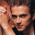 Pixelated image of a person with light brown hair and dark eyes, partially obscured by another figure's arm on the left side against a solid green background.