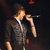 Pixelated image of a person singing into a microphone, wearing dark clothing with light accents against a dark background.