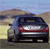 A dark-colored sedan viewed from the rear, positioned on a road with a blurred background of sky and terrain.