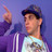 Pixelated image of a person in profile wearing a dark cap and light shirt, with a raised hand and microphone against a purple background.