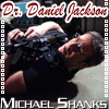 A man in a black shirt lies on his back with arms raised, holding a dark fabric over his face, under text reading "Dr. Daniel Jackson" and "Michael Shanks".