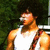 A person in a white tank top with a red and brown strap over the shoulder, standing against green foliage background.
