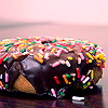 A chocolate-glazed donut with multicolored sprinkles on a reflective surface under soft lighting.