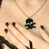 A close-up of a person's chest with a black skull and crossbones pendant on a silver chain necklace, fingers with dark nail polish visible.