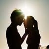 Silhouetted couple in profile facing each other, hands touching cheeks, backlit by bright sun creating lens flare.