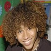 A person with voluminous curly brown hair wearing a dark top and layered necklaces, smiling against a blurred background with green and red elements.