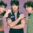 Three figures with dark hair stand against a purple background; the central figure wears a dark sleeveless top with light trim and has arms crossed.