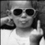 Black-and-white image of a person wearing sunglasses and a white shirt, making a middle finger gesture with the right hand while holding an object near the left ear.
