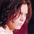 Pixelated image of a person with long brown hair wearing a dark red top against a dark background.