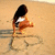 A person with long dark hair in a white top and brown shorts bends over on sandy ground, drawing or writing in the sand.