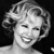 Black-and-white pixelated image of a smiling woman with styled hair, cropped to show shoulders and head against a dark background.