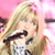 Pixelated image of a person with long blonde hair holding a black microphone against a pink background.